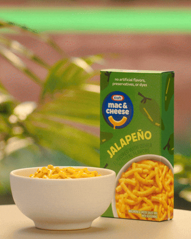 A bowl of Kraft Mac & Cheese Jalapeño flavor next to its green packaging box on a white surface with a blurred outdoor background.