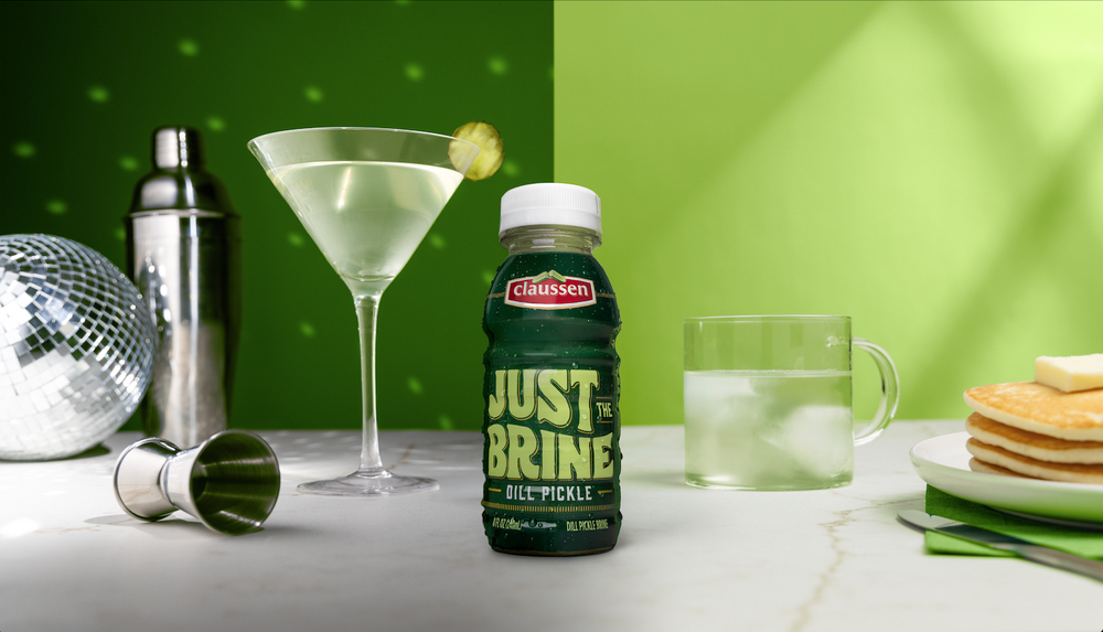 Various drink-related items on a table, including a cocktail glass with olives, a bottle of Claussen Just the Brine oil pickle, a glass of water with ice, and a green background with a disco ball and stacked pancakes on a plate.