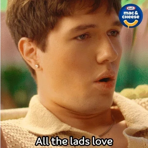 Close-up of a young man with short brown hair and earrings, looking surprised or intrigued, in a scene from a TV show or movie. There is a Kraft Mac & Cheese logo in the top right corner. Caption reads 'All the lads love'.