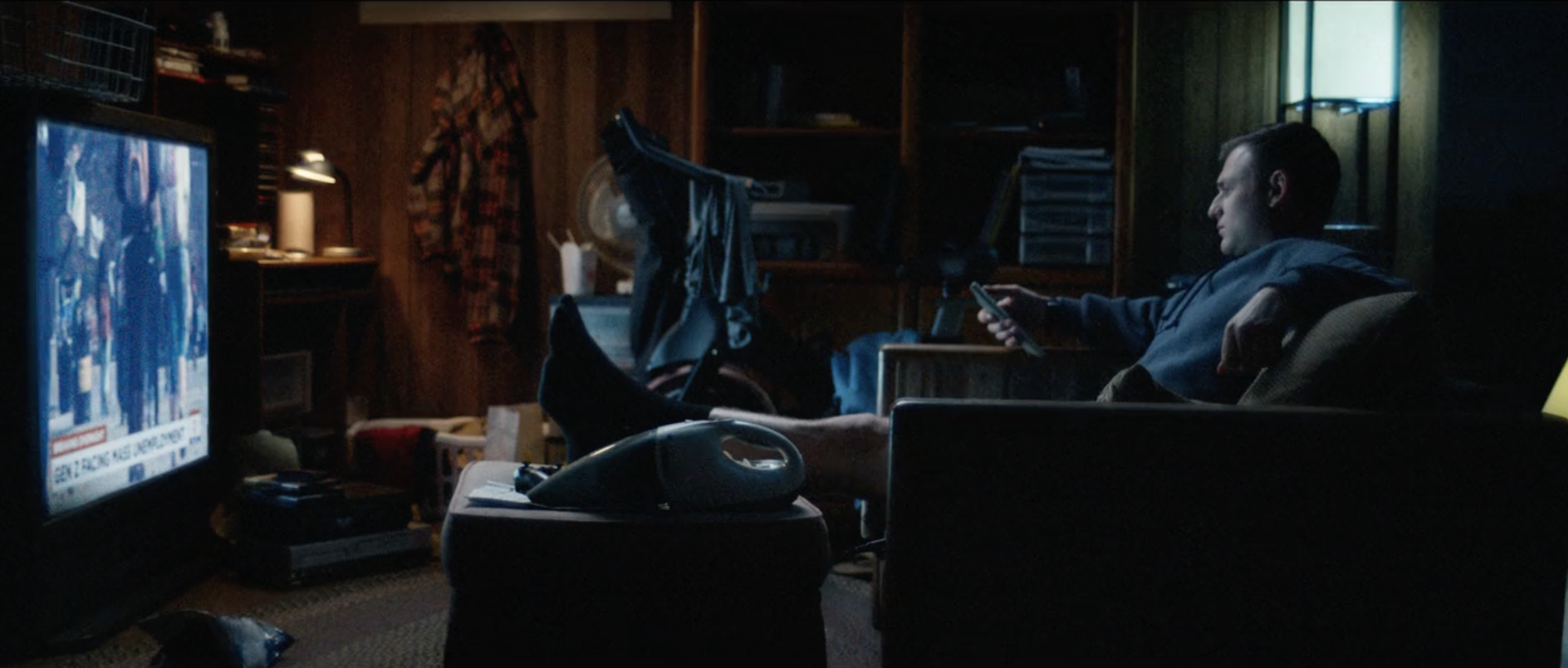 A man sitting on a couch in a dimly lit room, watching TV and using his phone, with a baby stroller nearby and cluttered surroundings.