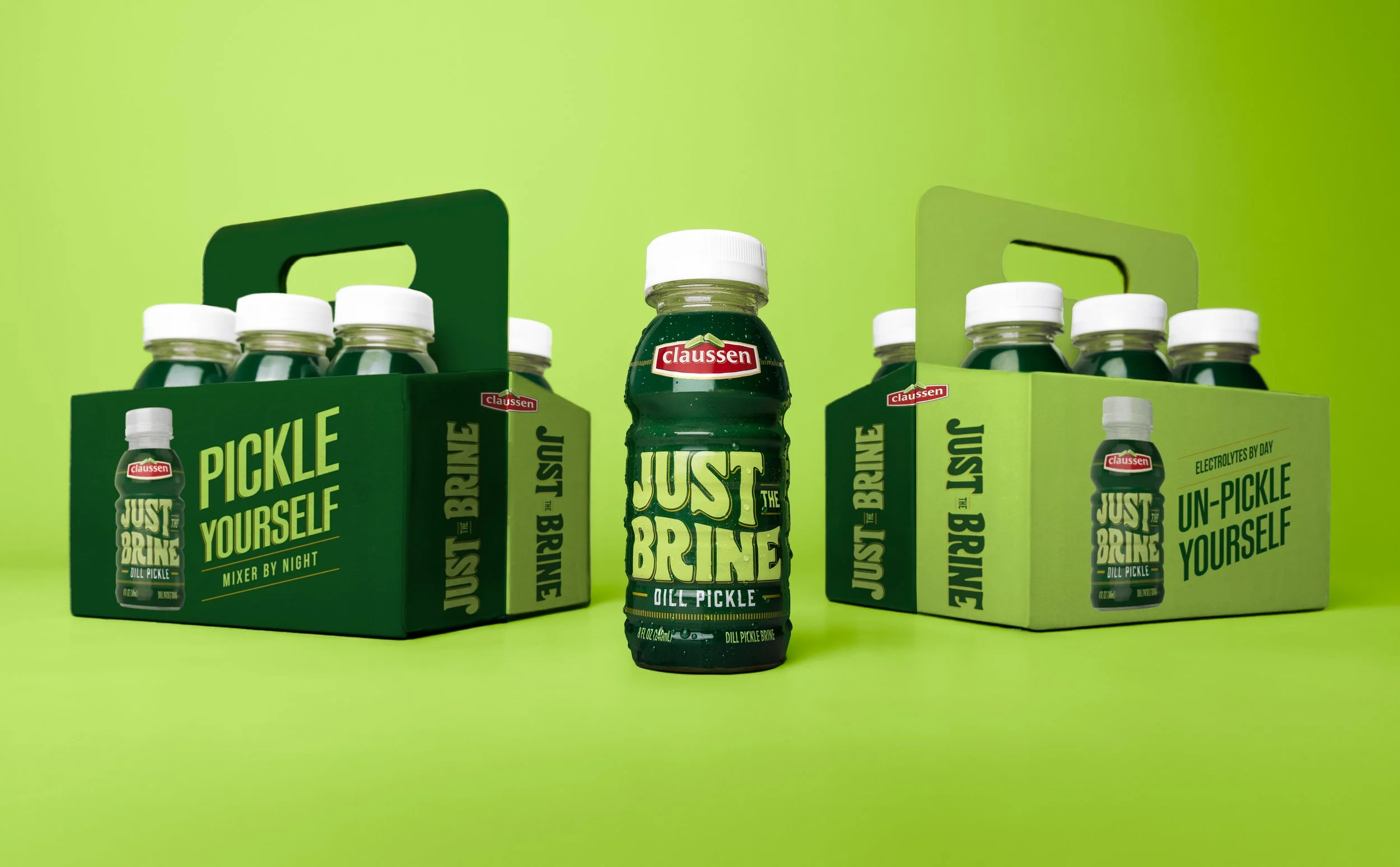 Center of the image shows a bottle of Claussen Just the Brine Dill Pickle with a white cap. Flanking it on both sides are green boxes each holding six bottles of the same pickle products in green bottles with white caps. The background and surface ar