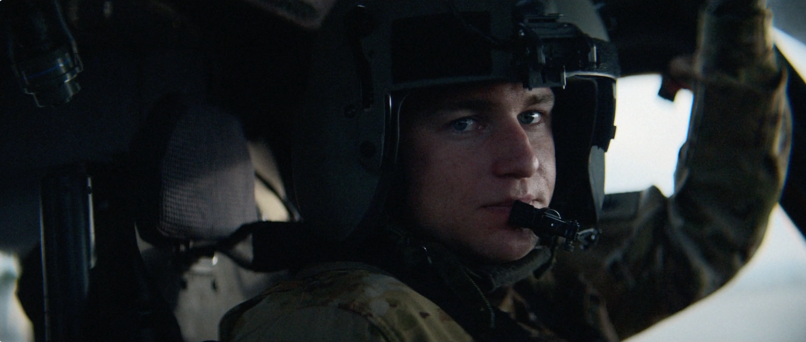 Close-up of a soldier inside a military helicopter cockpit, wearing a helmet with a mic and tactical gear, looking out.