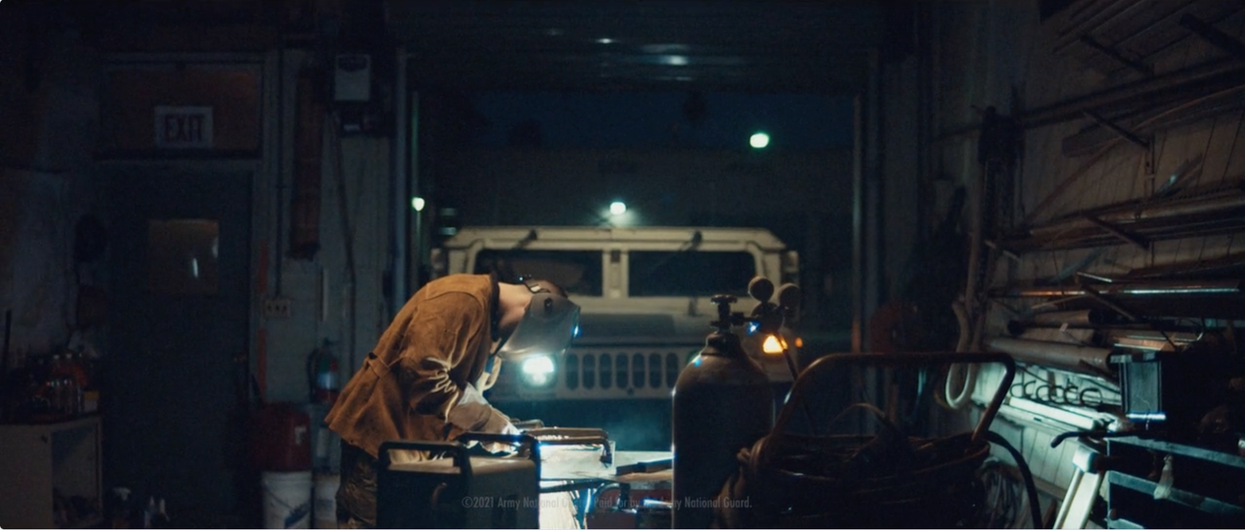 A person wearing a protective face shield and a brown jacket is working on a project at a workbench in a dimly lit garage or warehouse. There are various tools and equipment, including gas cylinders and a vehicle in the background.