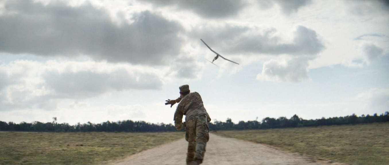 A soldier in camouflage uniform running on a dirt road while a drone flies overhead in a cloudy sky.