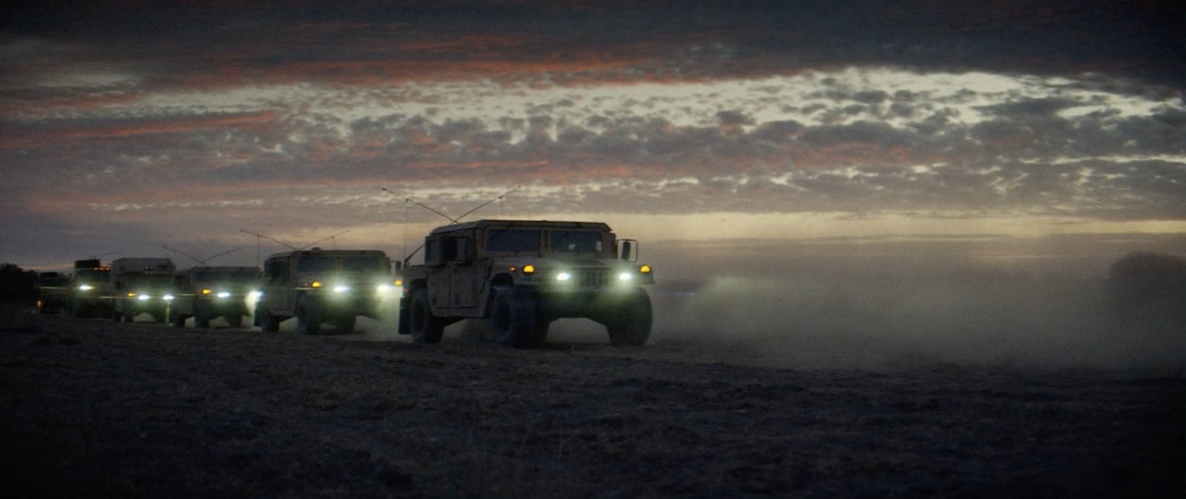 A convoy of military vehicles with headlights on, driving on a dusty road at dusk or dawn under a partly cloudy sky.