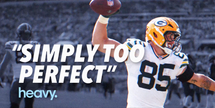 A Green Bay Packers football player wearing a yellow helmet with a white and blue uniform, holding a football in the air, celebrating during a game with blurred stadium background.