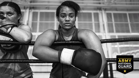 Two female National Guard soldiers wearing boxing gear, standing behind the ropes in a training gym. They look directly to camera.