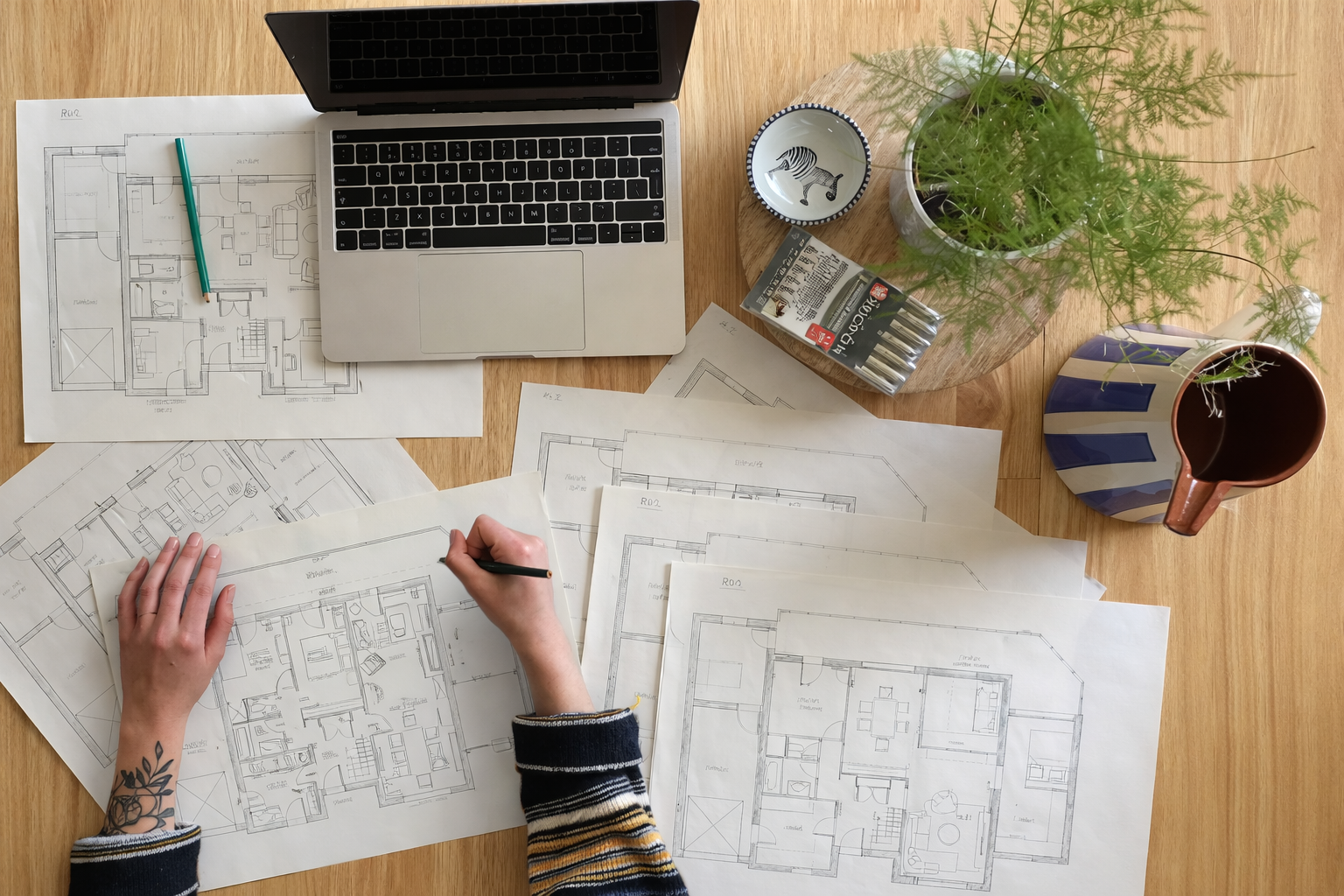 Plans d'architecture étalés sur une table, une personne les examinant avec un ordinateur portable ouvert, des plantes en pot, une tasse décorée de rayures, un petit bol avec un zèbre illustré, un paquet de rouleaux de papier de dessin.