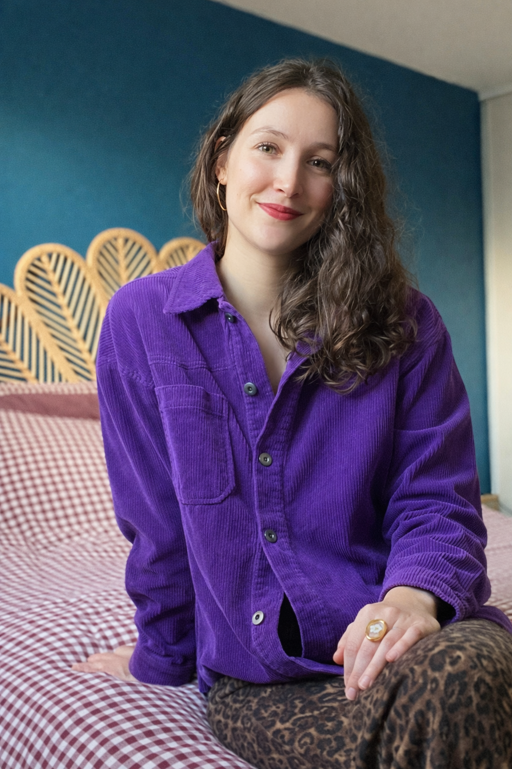 Jeune femme aux cheveux bouclés portant une chemise violette et un pantalon léopard, assise sur un lit avec un motif vichy rouge et blanc, dans une chambre avec un mur bleu et un panneau en osier en arrière-plan.