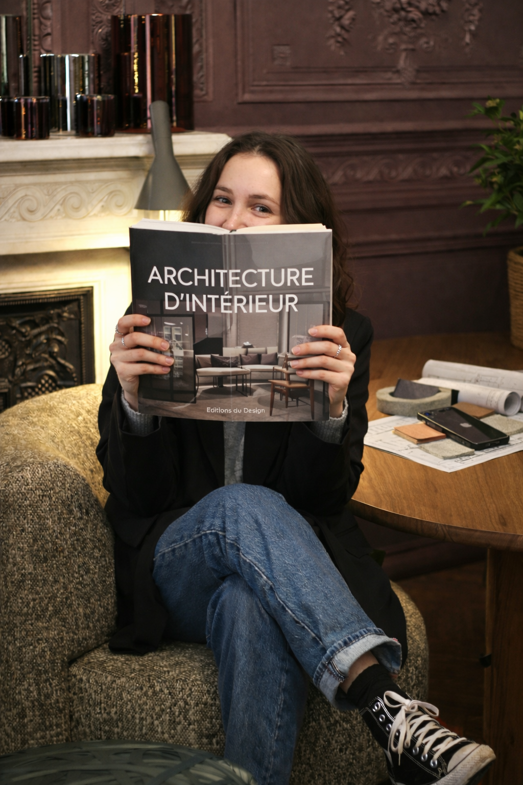 Jeune femme souriante tenant un livre intitulé 'Architecture d'intérieur' assis dans un salon sophistiqué avec une cheminée en marbre et une étagère remplie de livres.