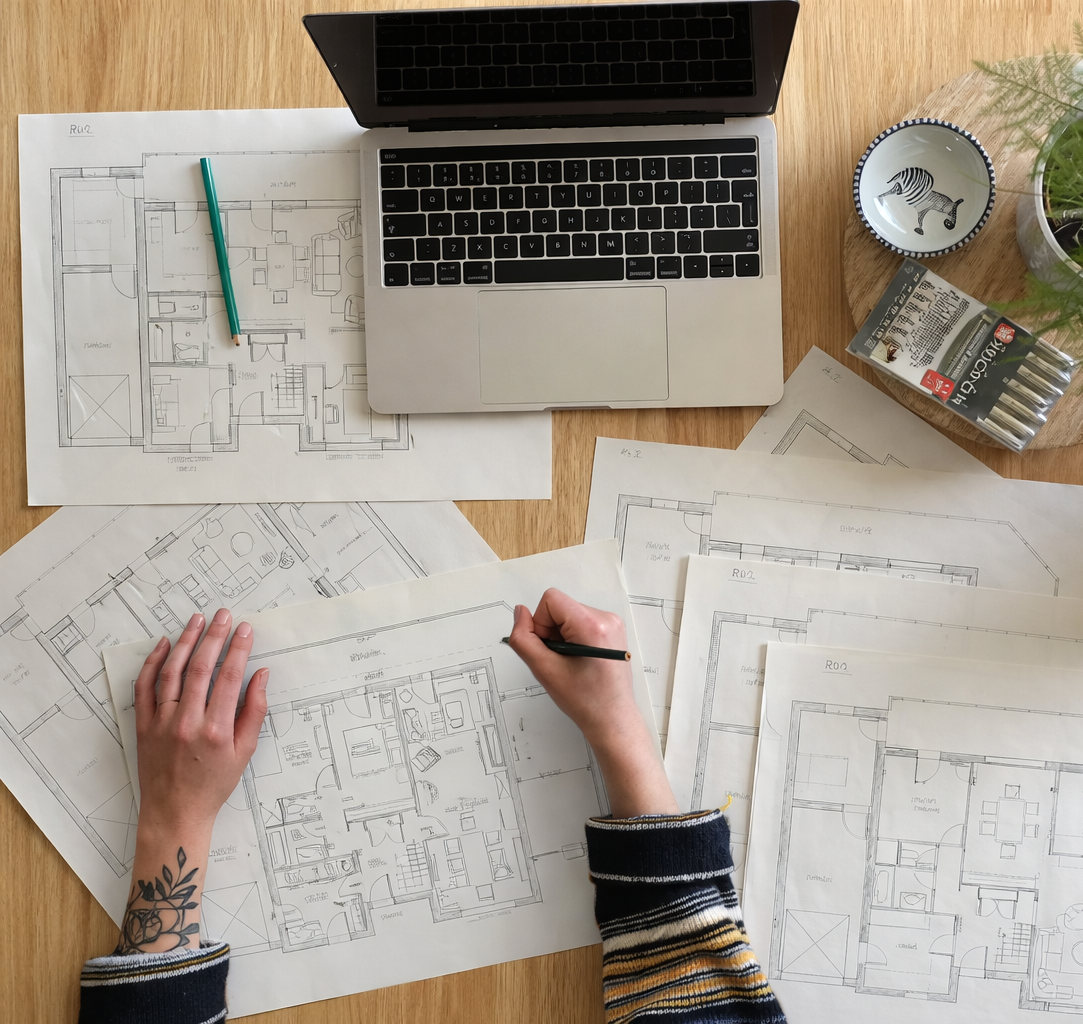 Une personne travaille sur des plans d'architecte de maisons, avec un ordinateur portable, des plans dispersés, une tasse à outil, une boîte de batteries, et une plante en pot.