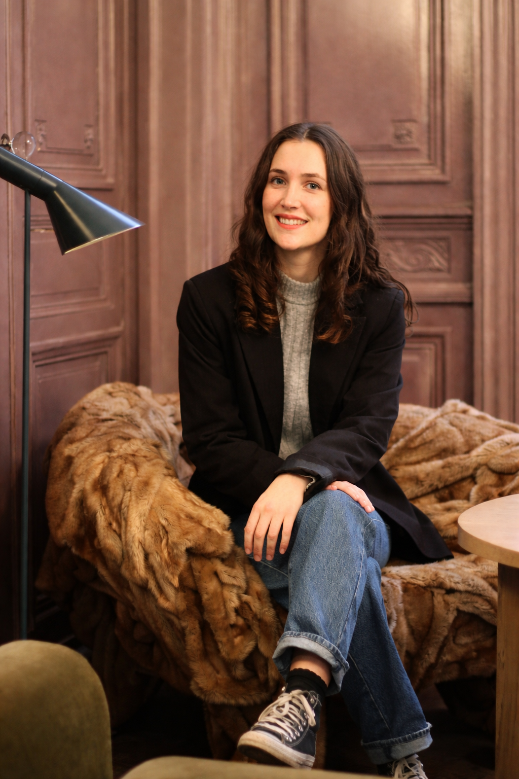 Une jeune femme assise sur un canapé en fourrure, dans une pièce avec des murs en bois, souriante, portant un blazer noir, un pull gris, un jean, et des Converse noires.