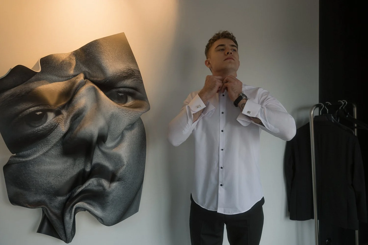 Young man in a white dress shirt adjusting his collar in a room with a large wall art sculpture of a human face.
