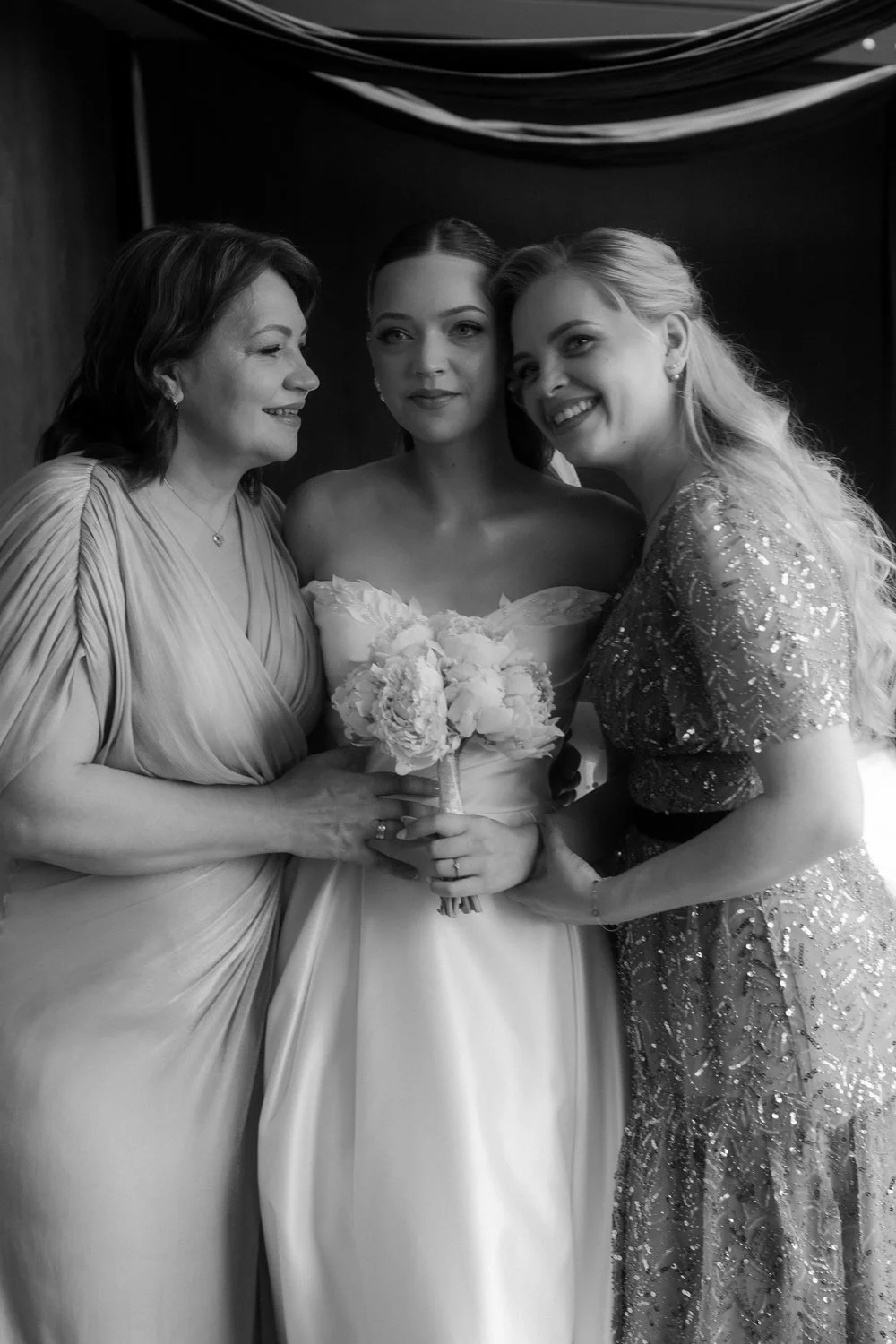 Three women, one in a wedding dress, standing together, smiling and holding a bouquet, at a wedding celebration.