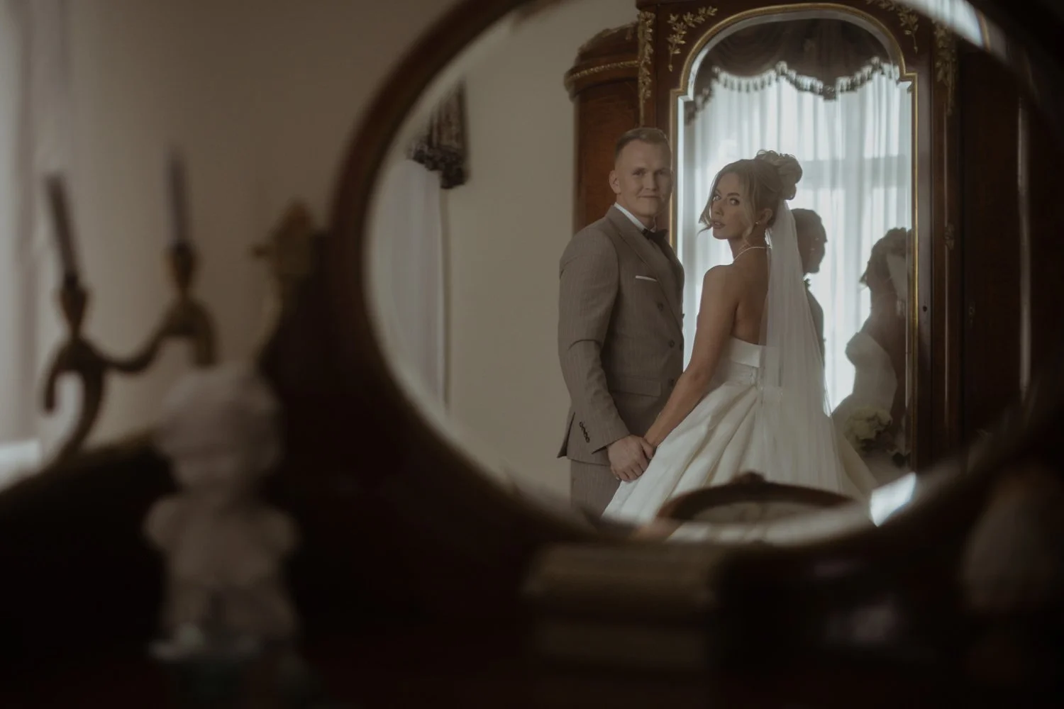 A bride and groom holding hands and looking at the camera are seen through a decorative vintage mirror, with a window with curtains in the background.