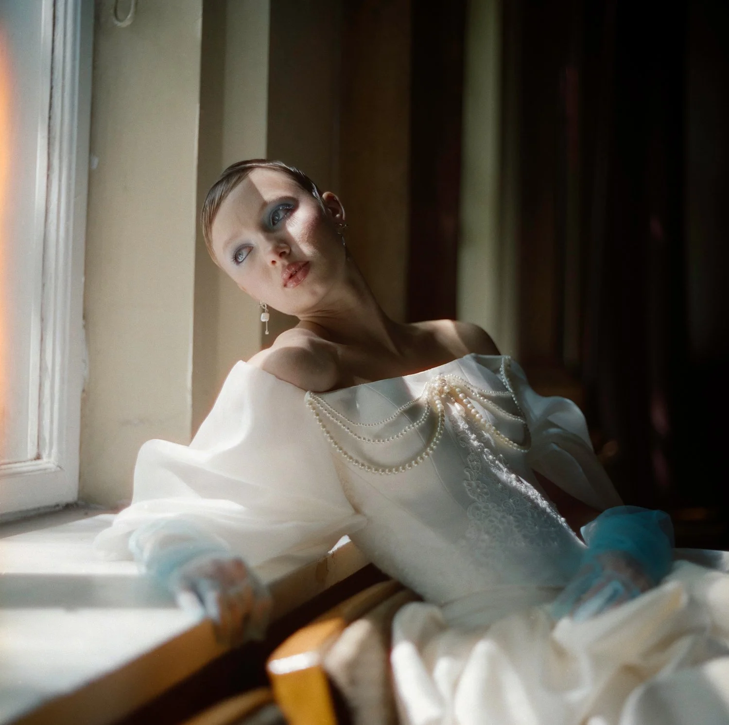 A woman with short hair and makeup sitting by a window, wearing a white dress with pearl embellishments and earrings, looking away from the camera in natural light.