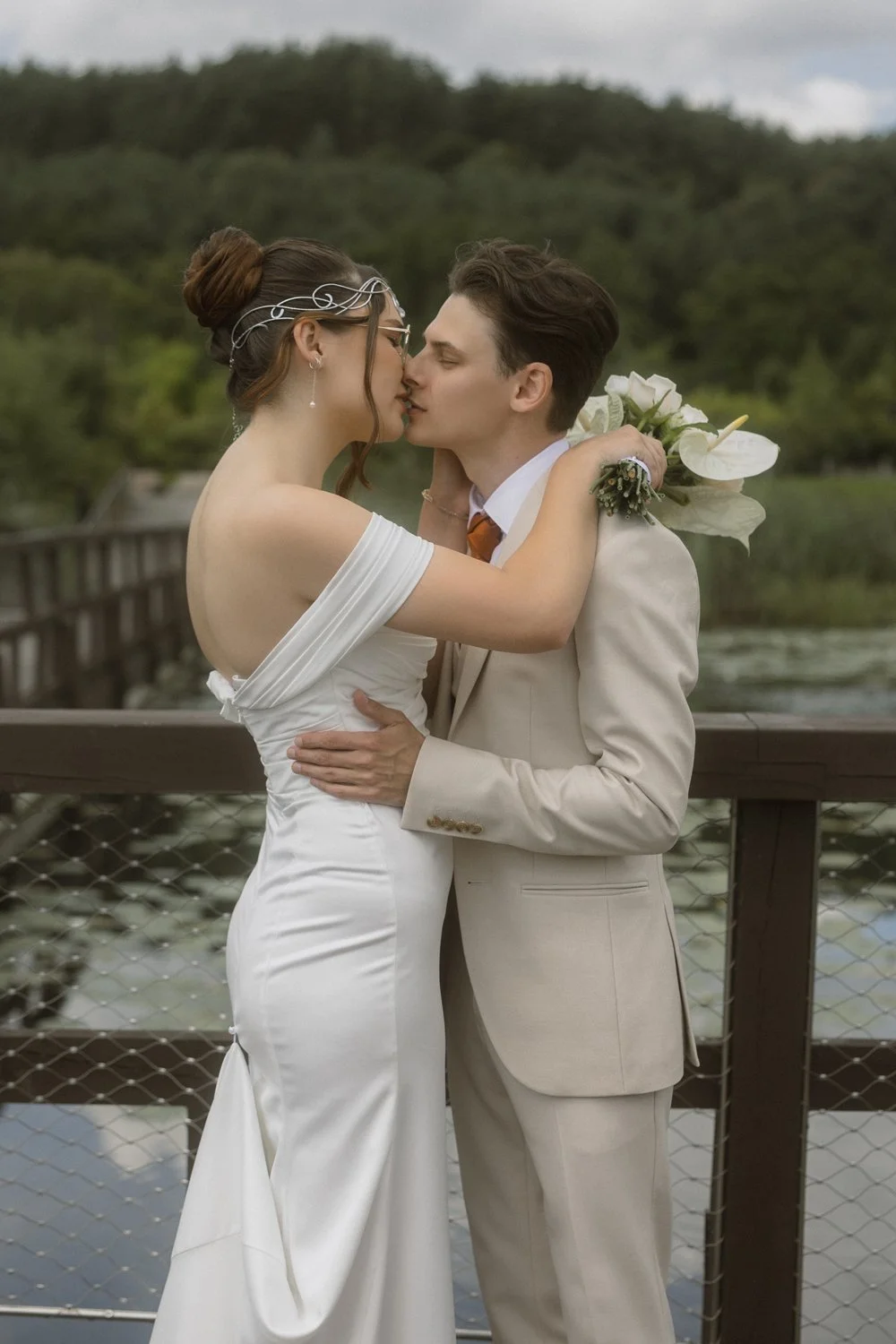 A bride and groom sharing a kiss on a wooden bridge near a lake, with a car and forest in the background.