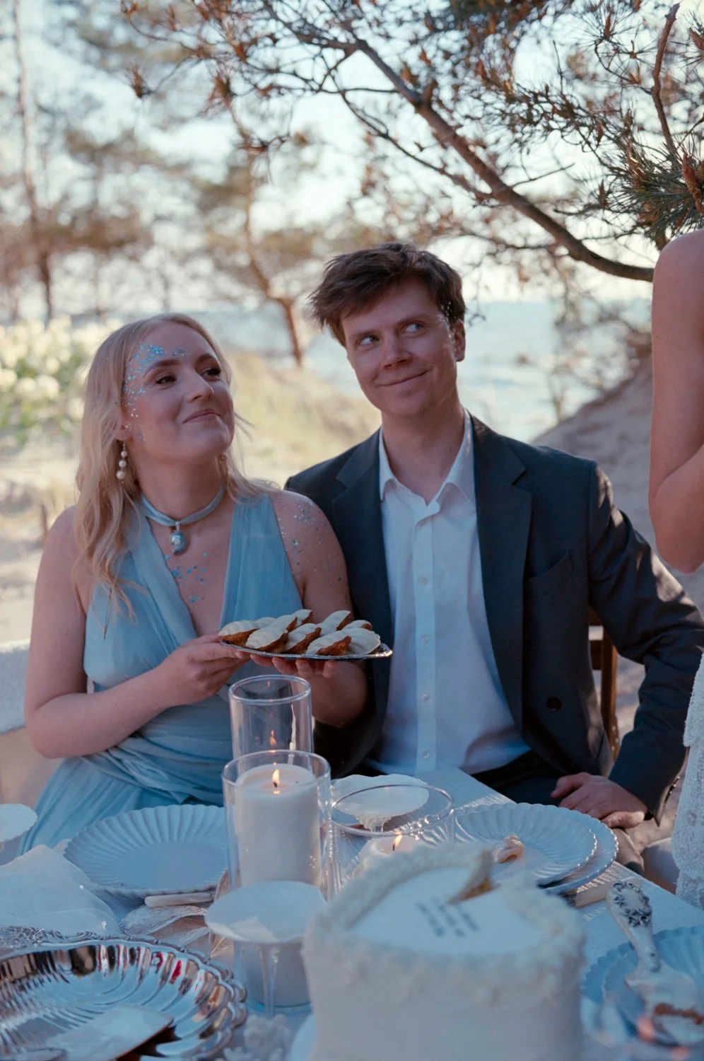 A woman dressed in blue with glitter on her face holding a plate of cookies, sitting beside a young man in a suit at an outdoor table set with candles, plates, and a cake, during a daytime gathering under trees.