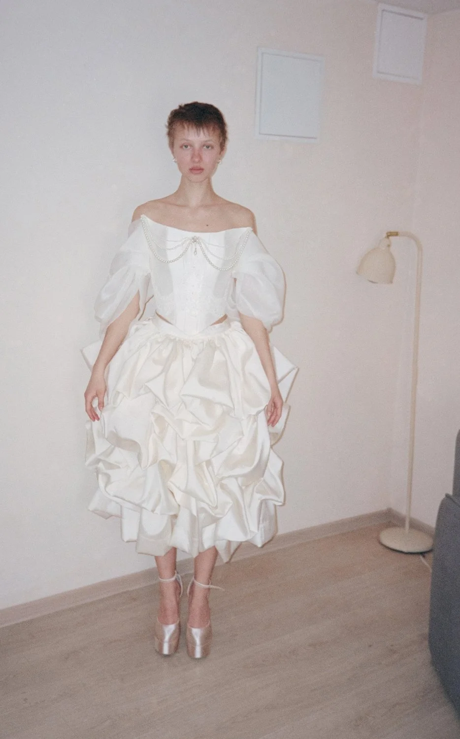 A woman standing indoors wearing an elegant, vintage white dress with puffy sleeves, ruffled skirt, and pearl embellishments, paired with high-heeled shoes.