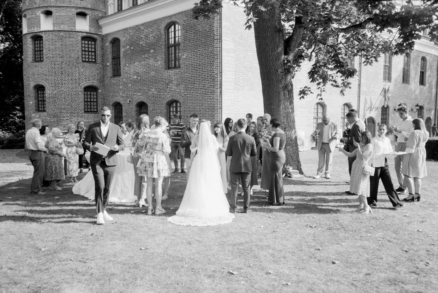 A black and white photo of a wedding reception outdoors next to a large brick building with several windows. The bride, wearing a veil and white gown, and the groom, in a suit, are at the center talking to a group of people. Guests are mingling, some