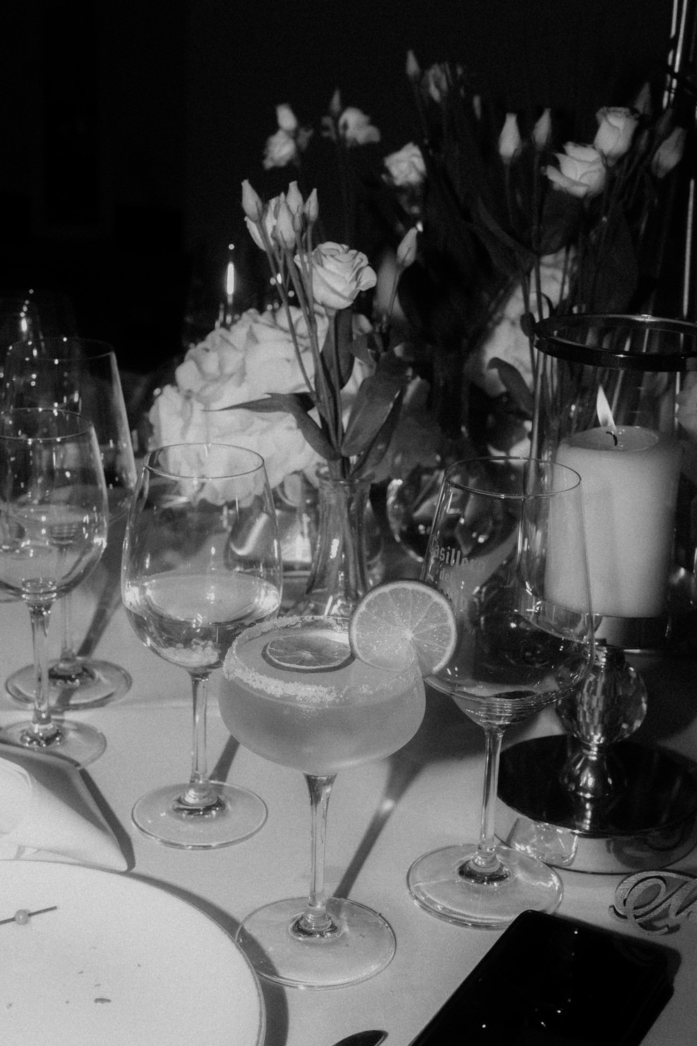 A table set with wine glasses, a cocktail with a lemon slice, a bouquet of roses, and candles in a dimly lit setting.