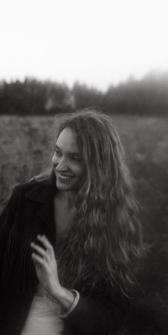 A woman with long, curly hair is smiling and looking to her left outdoors during twilight or dusk, with a blurred landscape of trees and an open field in the background.