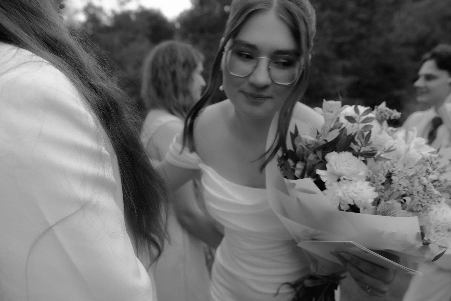 A woman in a wedding dress holding a bouquet of flowers, wearing glasses, smiling, with other people in the background at an outdoor wedding ceremony.