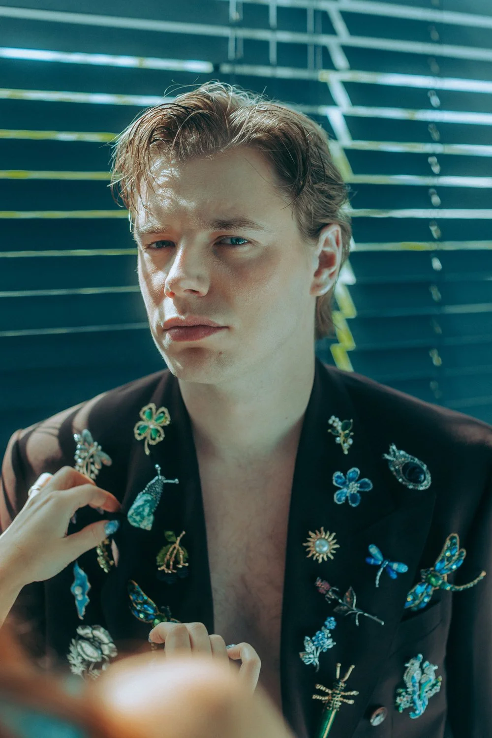 A young man with light skin and brown hair, styled back, sitting in front of blue blinds. He wears a black blazer decorated with colorful embroidered patches of insects, flowers, and other designs. A person’s hand is adjusting a pin on the blazer.