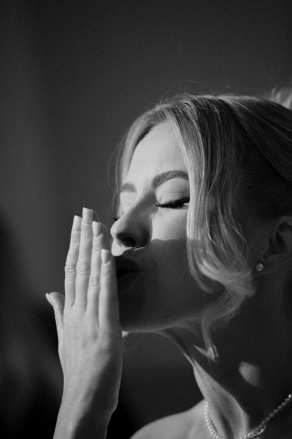 Black and white close-up of a woman with her eyes closed, touching her lips with her hand, wearing pearl earrings and a necklace.
