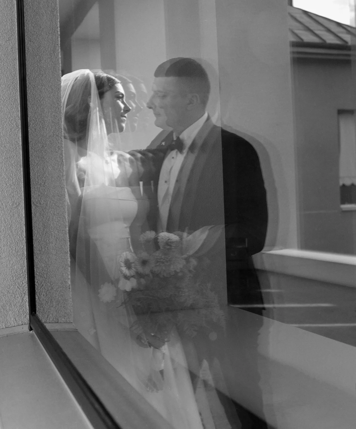 A black-and-white photo of a bride and groom seen through a glass window, smiling at each other. The bride holds a bouquet of flowers, and they are dressed in wedding attire.