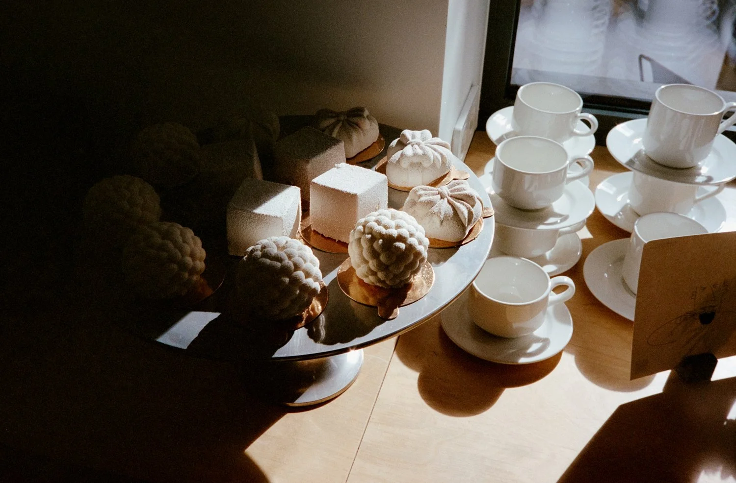 A display of various white and beige desserts on a silver tray and a table with white coffee cups and saucers near a window.