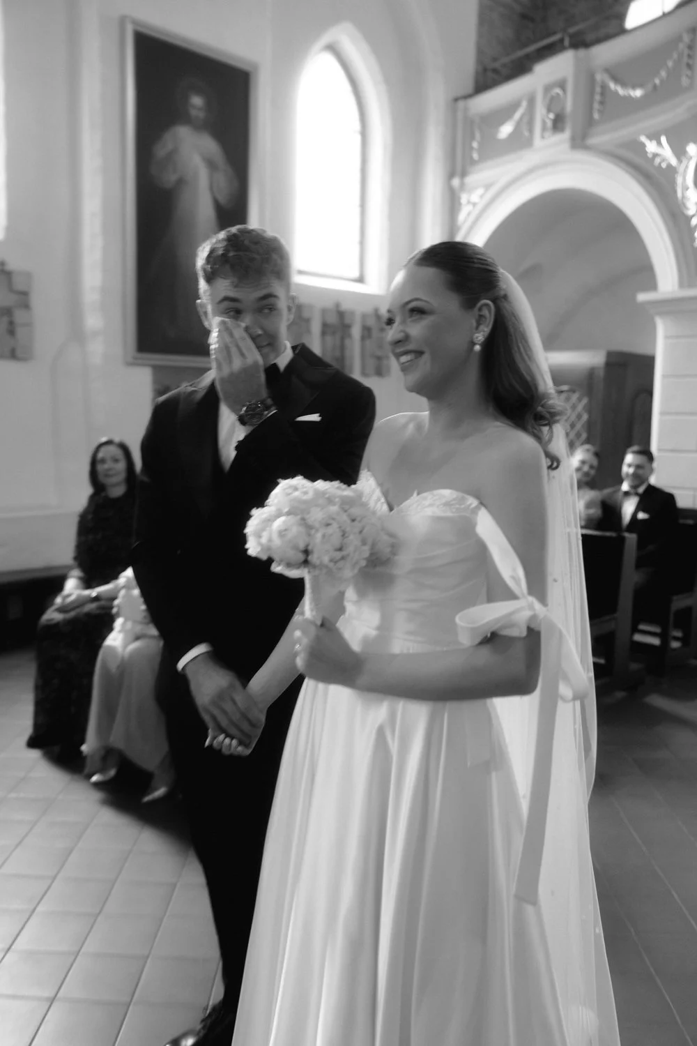 A bride and groom standing inside a church during their wedding ceremony, with the groom wiping a tear from his eye, holding the bride's hand, and the bride smiling holding a bouquet of flowers.