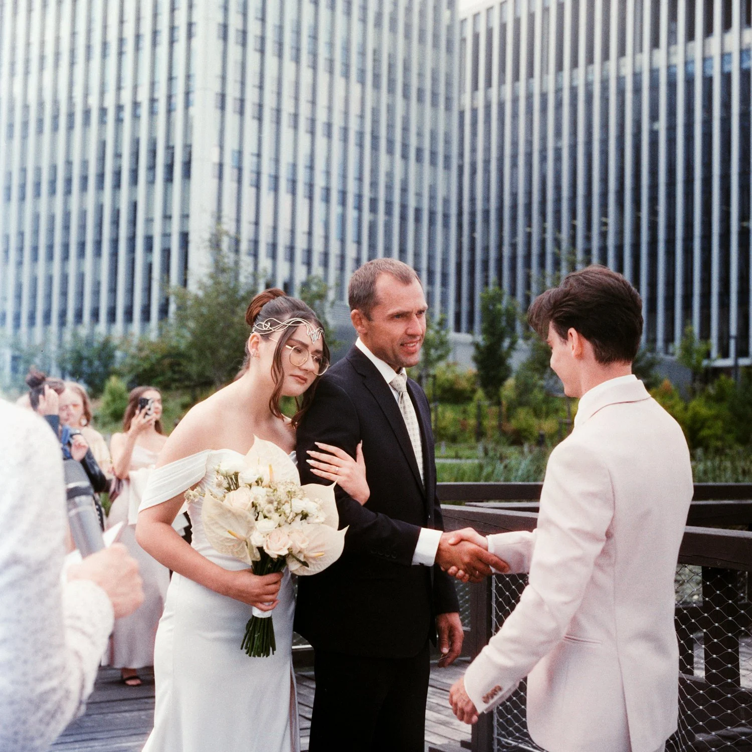 A wedding ceremony taking place outdoors in front of a modern building, with a bride and groom exchanging vows, the bride holding a bouquet and wearing a white dress, and the officiant shaking hands with the groom.