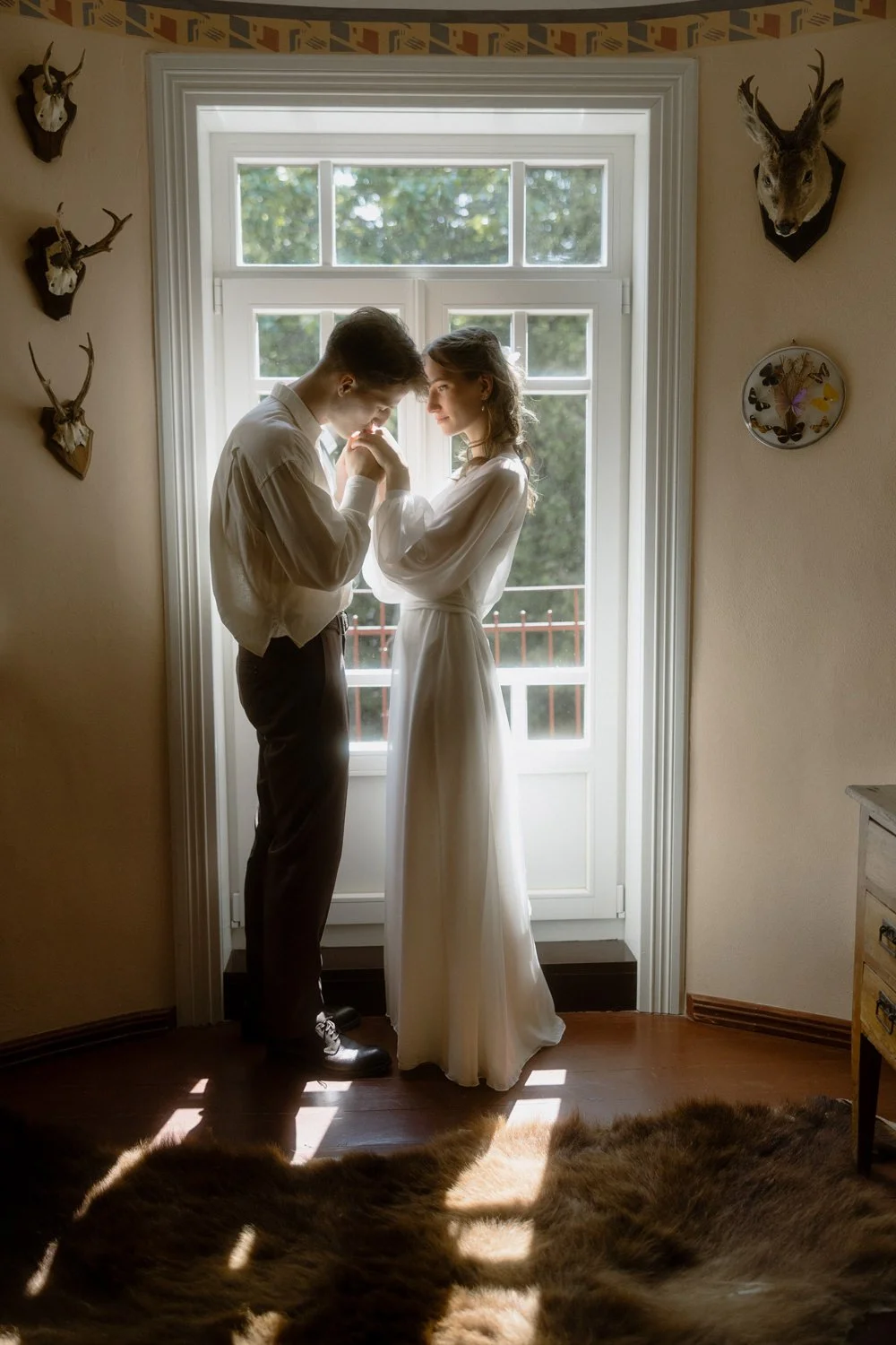 A couple standing close together inside a room with a large window, backlit by natural light, with decorative deer head mounts on the wall and a plush fur rug on the wooden floor.