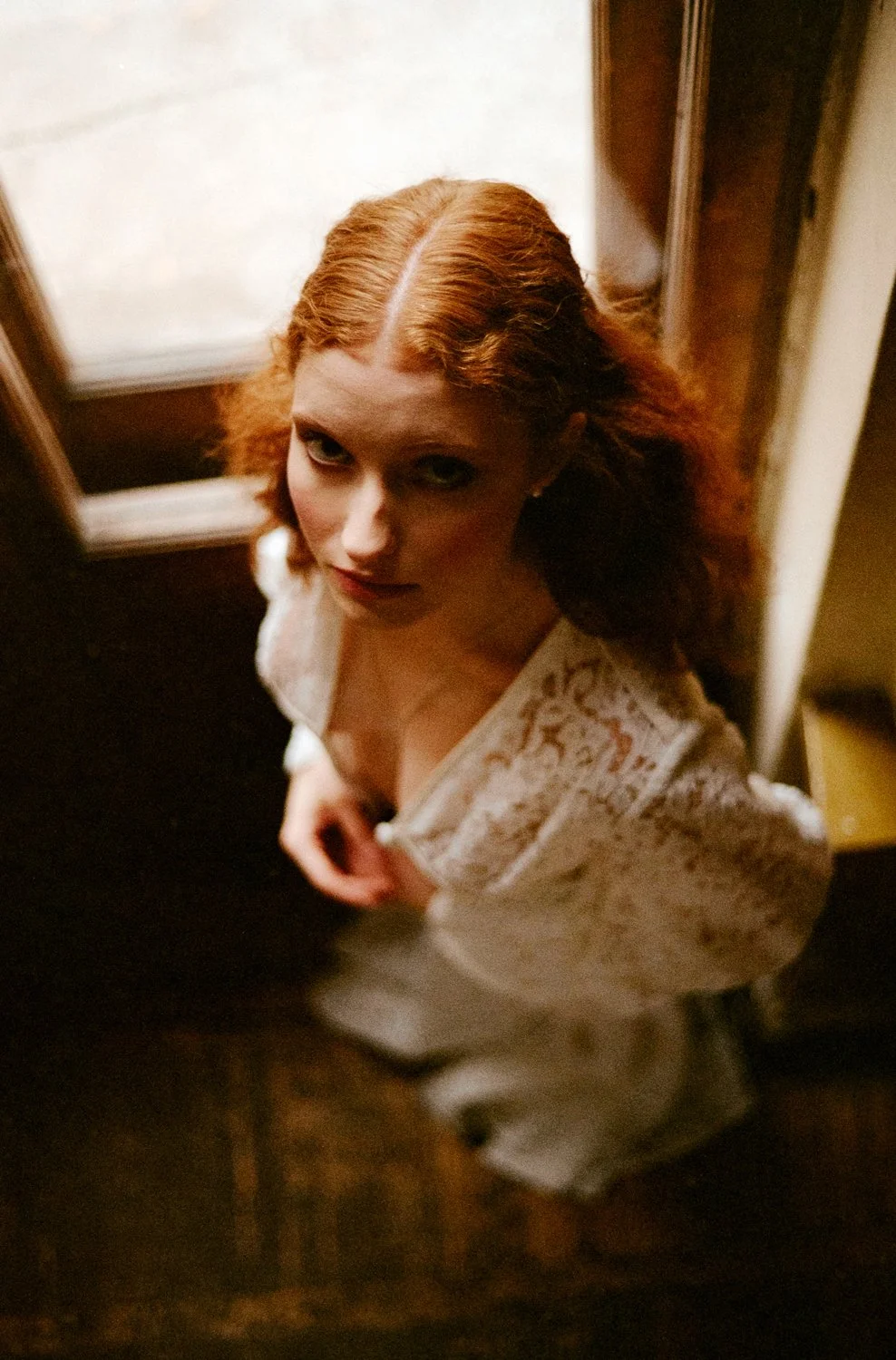 A woman with red hair looking up at the camera, standing near a window with wooden framing, wearing a white lace top.