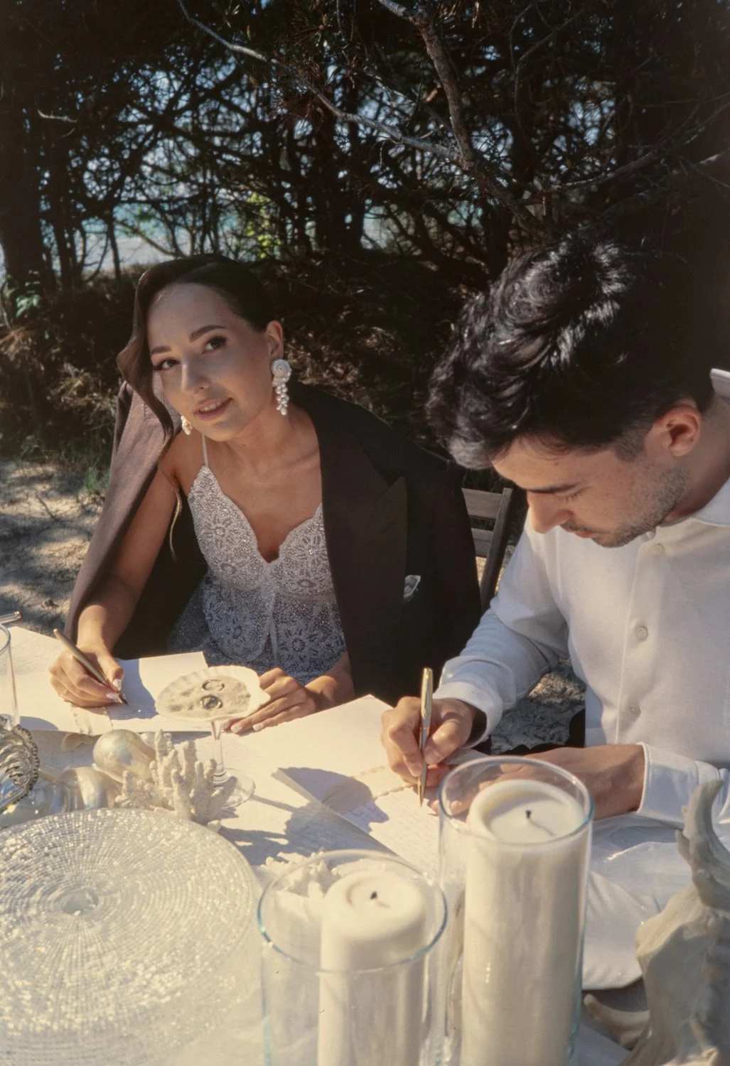 A woman in a black blazer and white lace dress with large earrings, sitting at a table outside, signing a document, with a man in a white shirt also signing a paper, surrounded by white candles and decorative items, with trees in the background.
