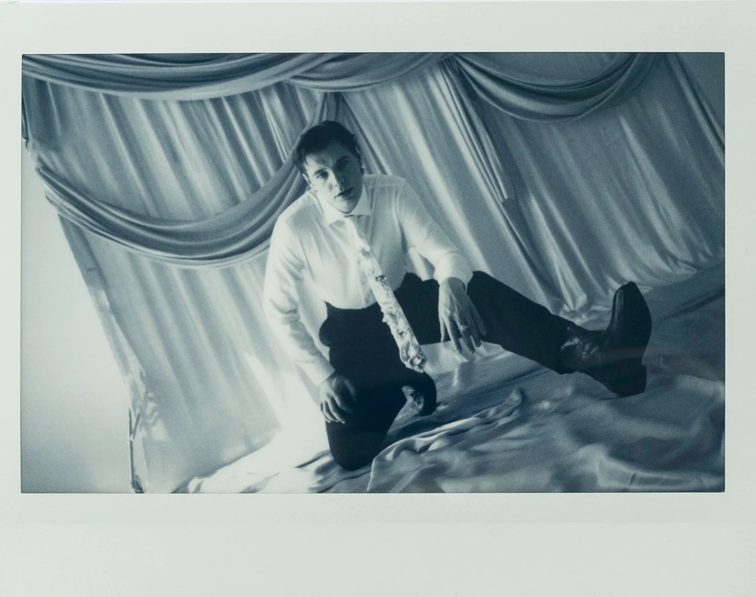 A person with dark hair, wearing a white shirt, a patterned tie, and black pants, is kneeling on a satin bed in front of satin curtains. The image is in black and white.