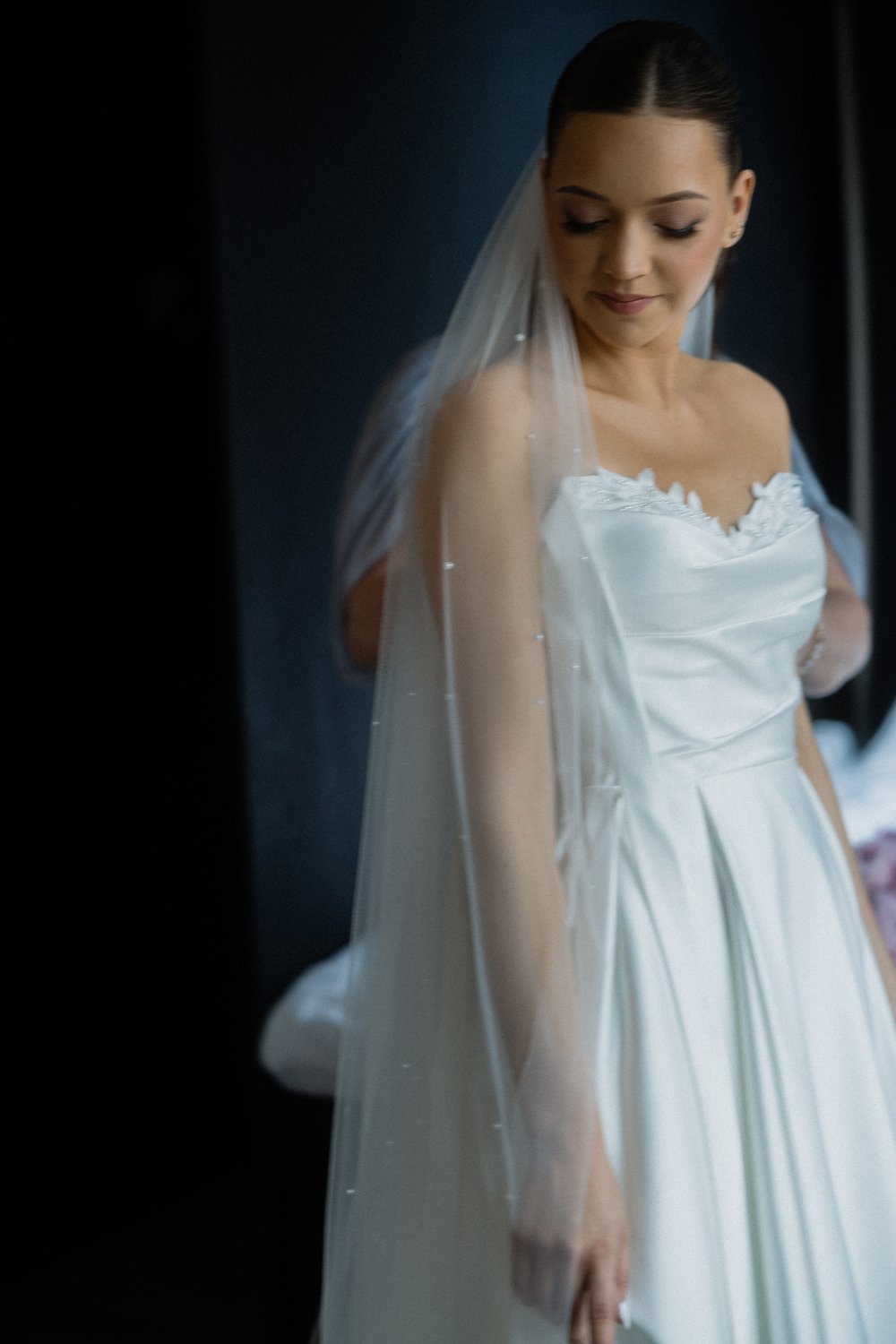 A bride in a white wedding dress and veil stands with her eyes closed and head slightly tilted downward, smiling softly.