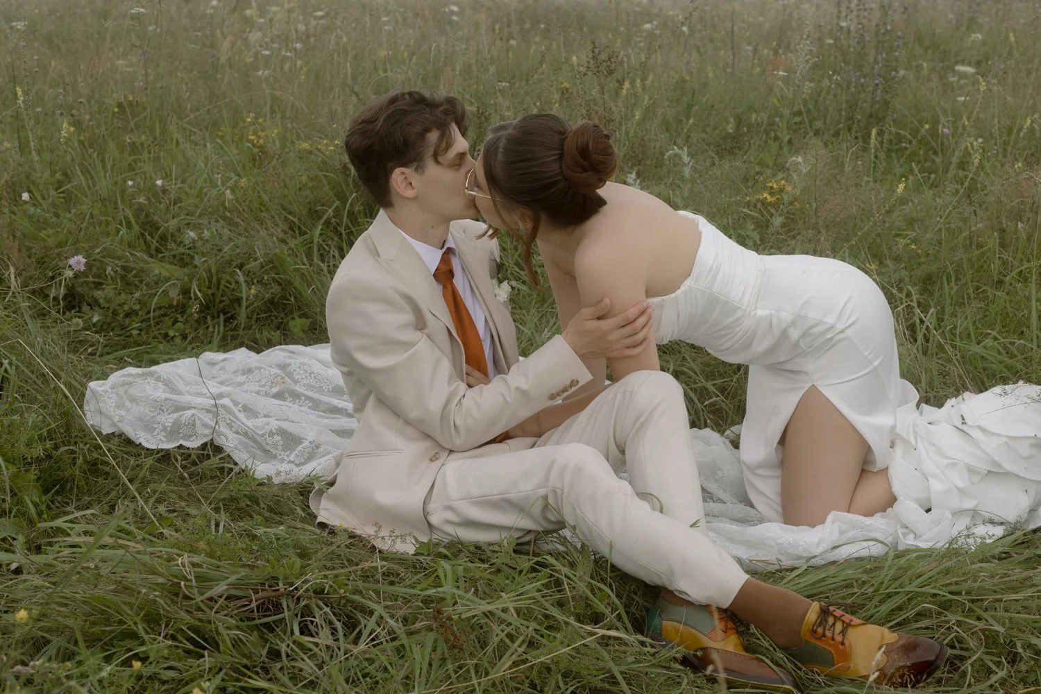 A man and woman sitting on a blanket in a grassy field, sharing a kiss. The man is dressed in a beige suit and orange tie, while the woman is wearing a white strapless dress. The woman is kneeling while the man is seated. The scene appears romantic a