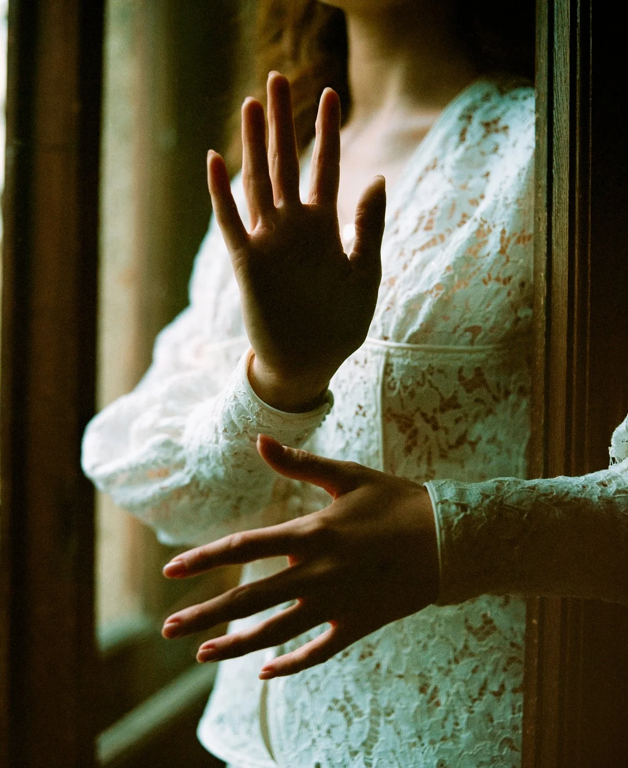 A woman in a white lace dress with puffed sleeves stands behind a window, touching the glass with her hand.
