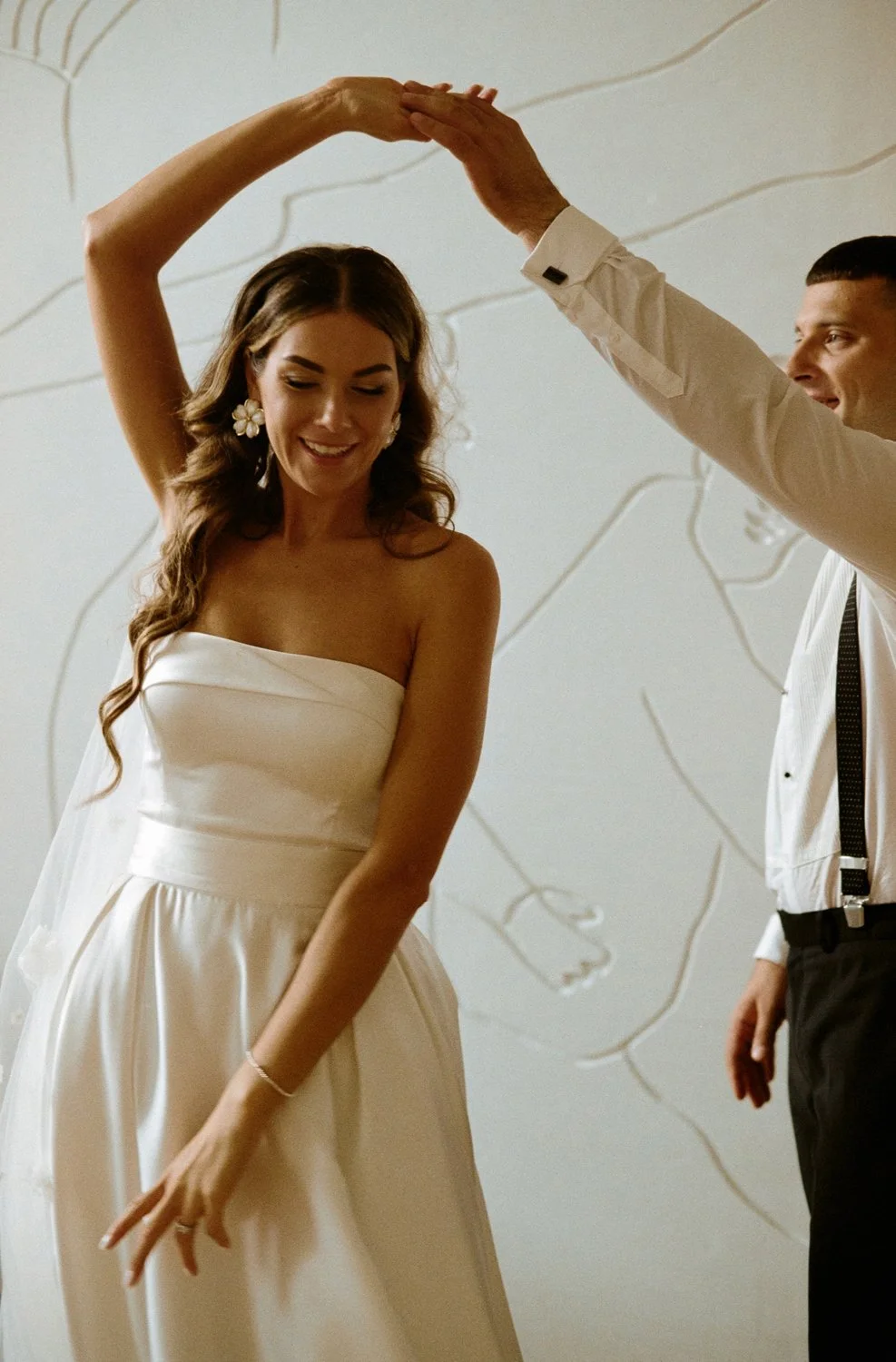 A woman in a strapless white dress dancing with a man in a white shirt and black suspenders, holding her hand overhead, with a light background.