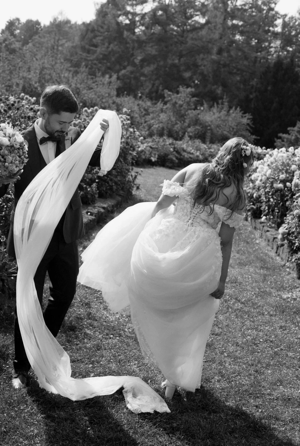 A newlywed couple outdoors, the groom is holding back a long tulle veil while the bride, in a wedding dress, adjusts her gown amid blooming bushes.