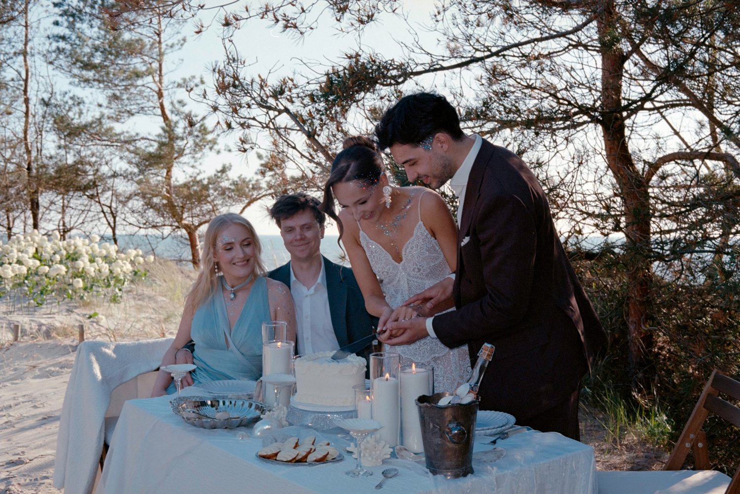 A wedding celebration taking place outdoors on a sandy beach during sunset with four people gathered around a table. Two women and two men are present, with the bride and groom cutting a wedding cake together while the others look on smiling.