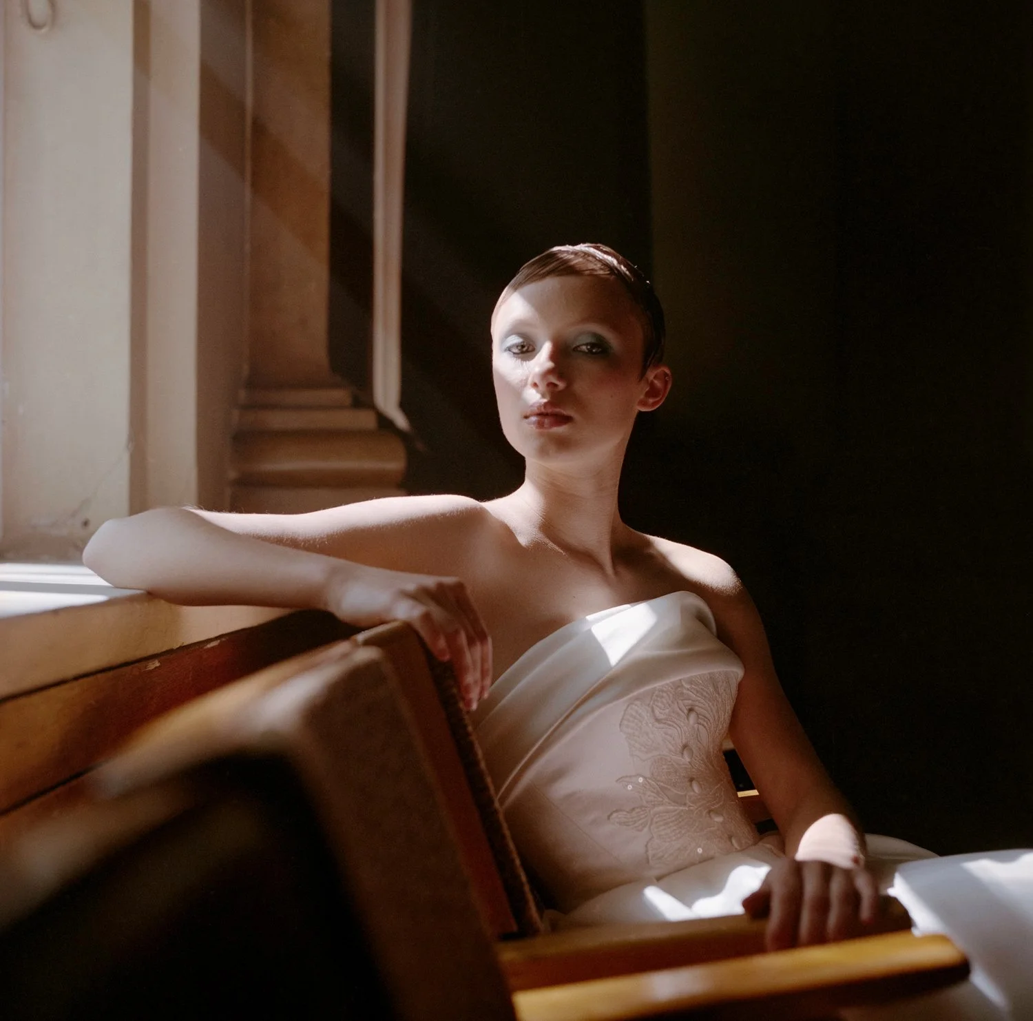 A woman with short dark hair and makeup, wearing a strapless white dress, sitting on a wooden bench in a dimly lit room with architectural details and a staircase in the background.