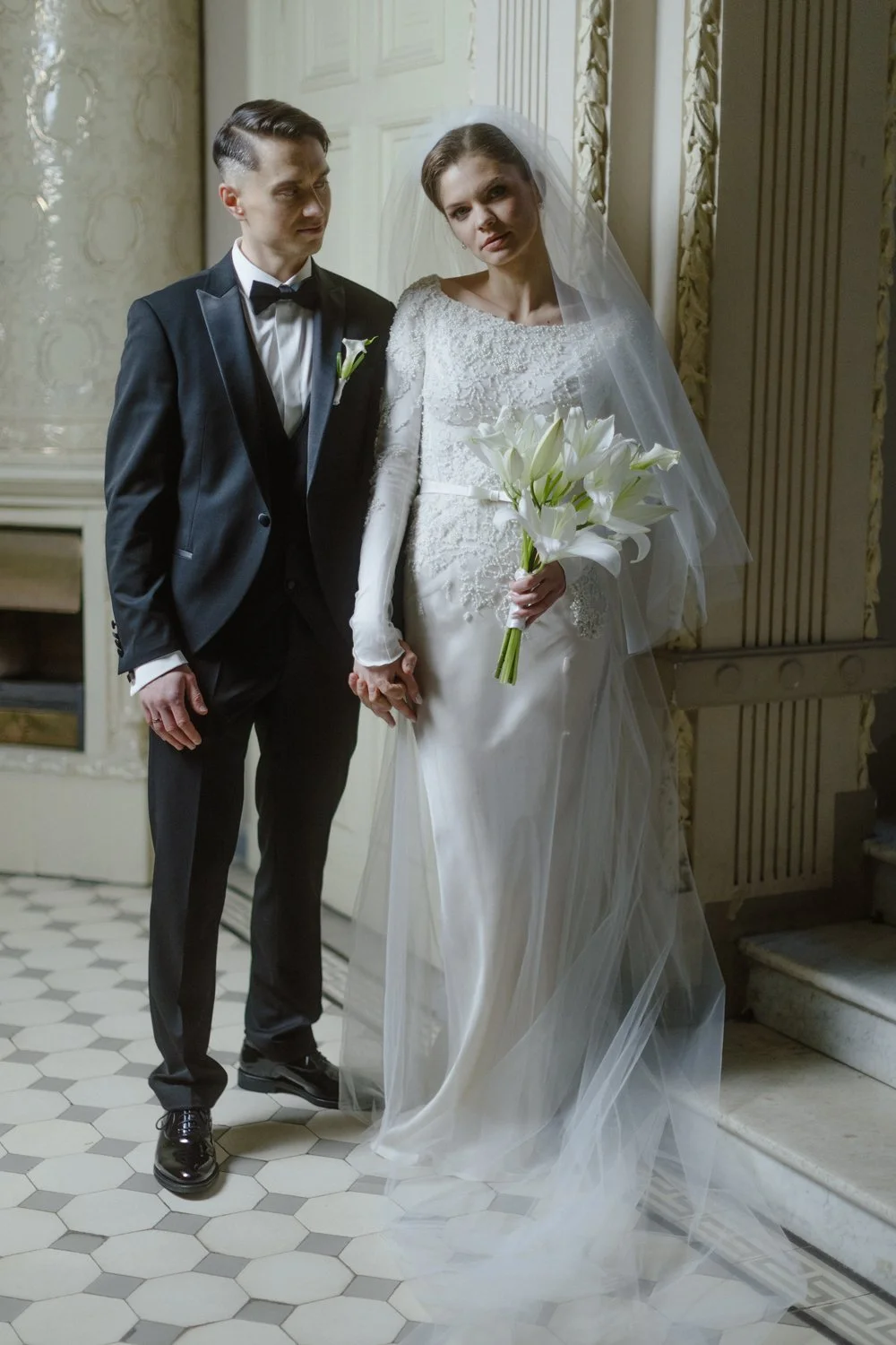Bride in a white wedding gown and veil holding a bouquet of white lilies, standing beside a groom in a black tuxedo with a bow tie, inside an ornate room with patterned walls and hexagonal tiled floor.