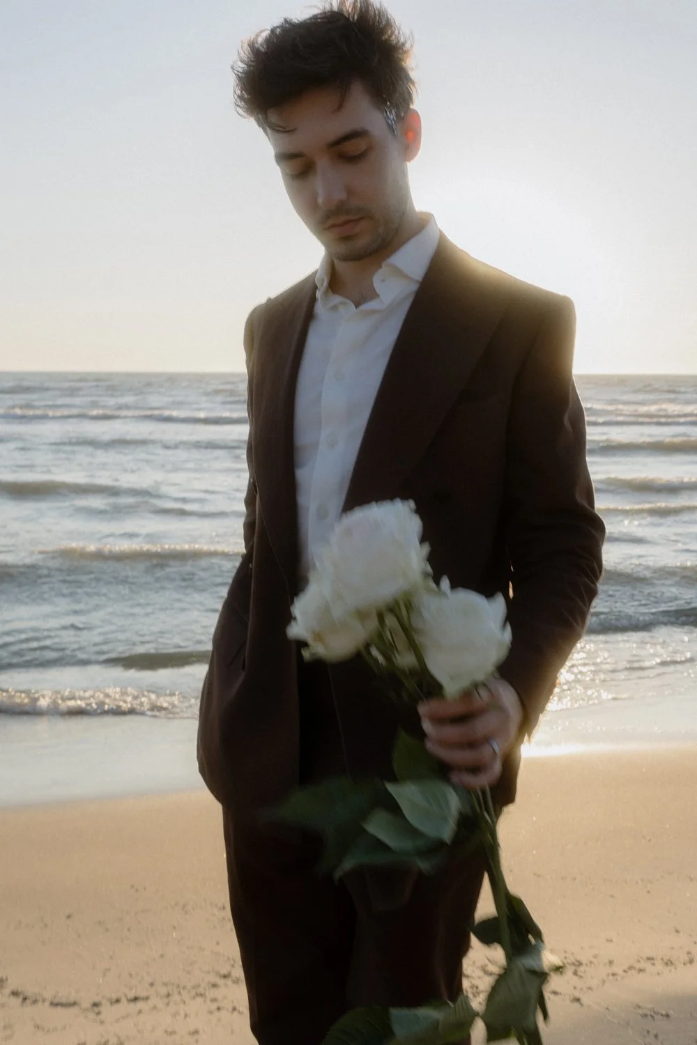 A man in a dark suit holding a bouquet of white roses on a beach at sunset.