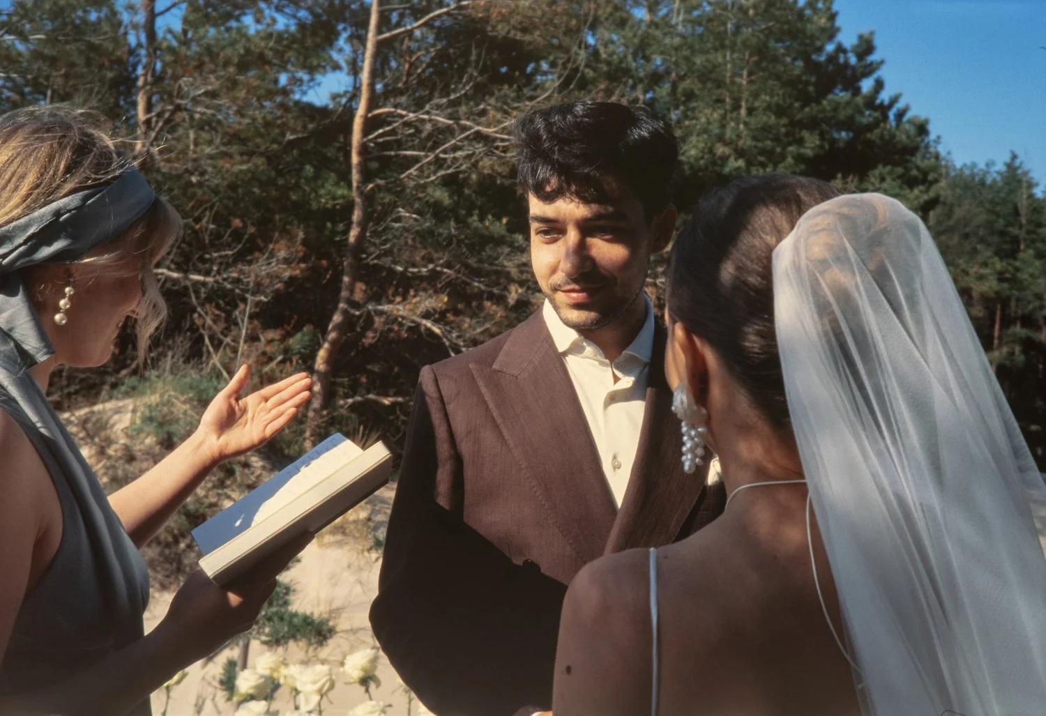 A couple getting married outdoors with an officiant reading from a book, under a clear blue sky and surrounded by trees.