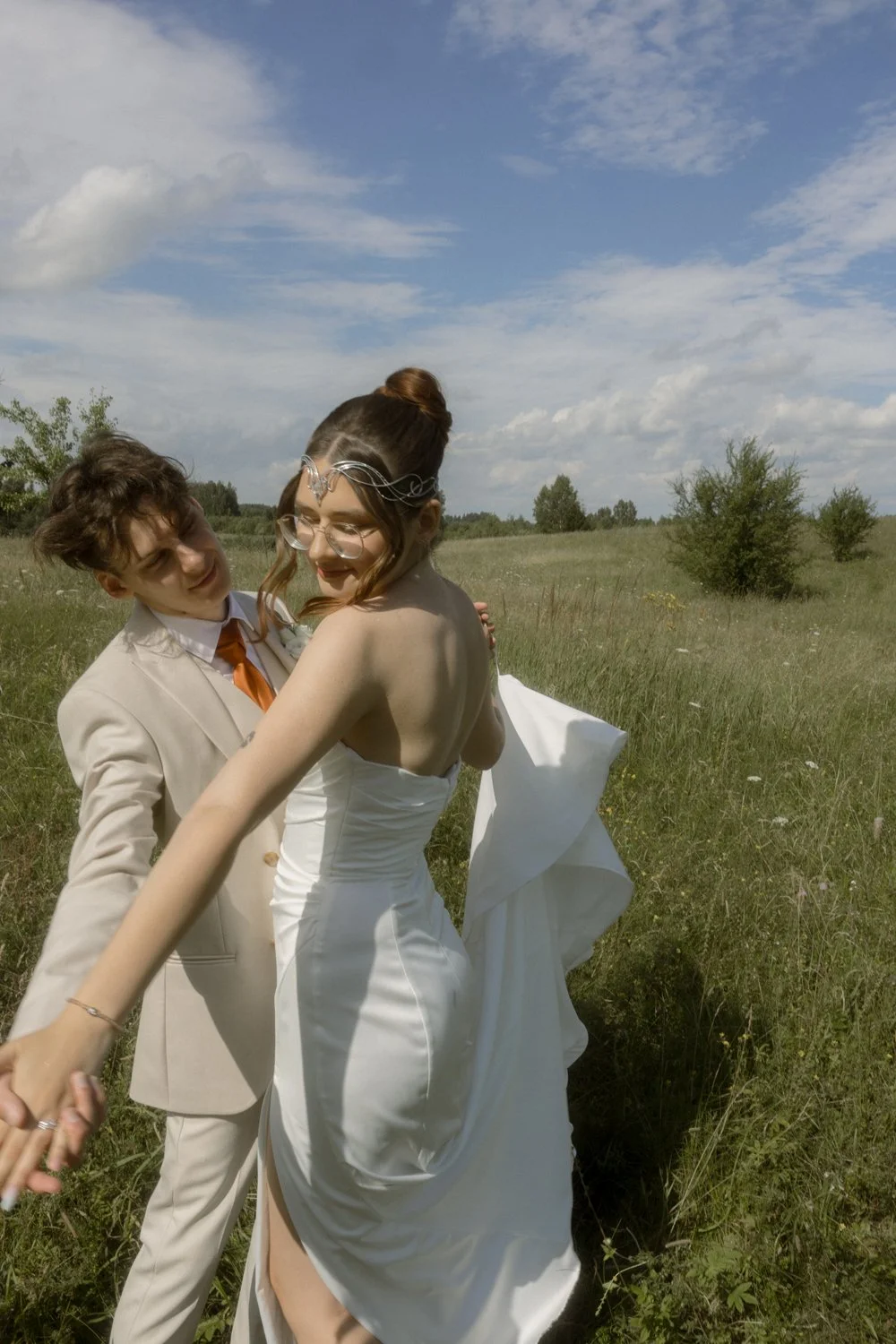 A couple dancing outdoors in a field on a sunny day, with the woman wearing a strapless white dress and the man in a light-colored suit with an orange tie.