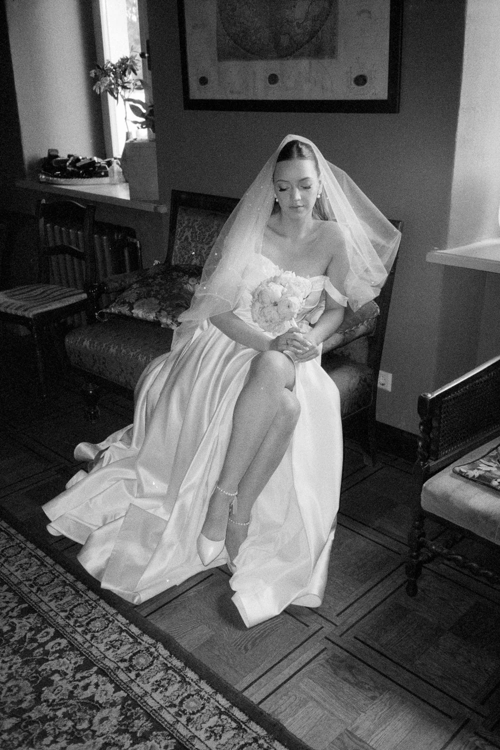 A bride sitting on a vintage sofa in a wedding dress with a veil, holding a bouquet of flowers, in a warmly decorated room.