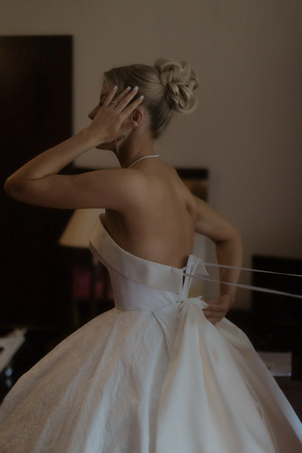 A woman in a strapless wedding dress with a full skirt and pearl necklace, adjusting her hair in a dimly lit room.